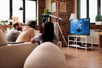 Playful casual multiracial friends sitting on sofa while playing space simulator videogame on gaming console. People in living room on sofa relaxing with games played on modern entertainment device.