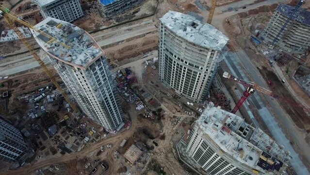Aerial view of a modern urban construction site, featuring multiple high-rise residential towers under development. Towering cranes lift materials. Earthworks, roads, and adjacent buildings. 
