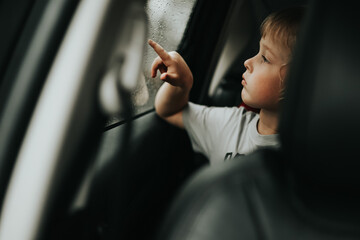 Thoughtful little blond baby boy seated in car backseat looking through rain covered window in rainy day.