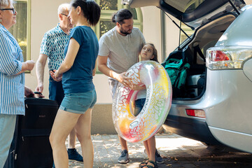 Parents with kid loading bags in trunk, putting baggage in car to leave on holiday vacation journey. Big family with grandparents travelling on summer trip to seaside with inflatable for leisure.