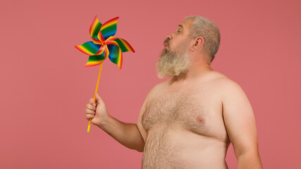 a gray-haired man with a naked torso blowing into a pinwheel. The spinner is painted in rainbow...