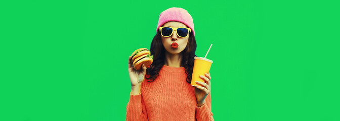 Portrait of stylish young woman drinking juice with burger fast food on green colorful background