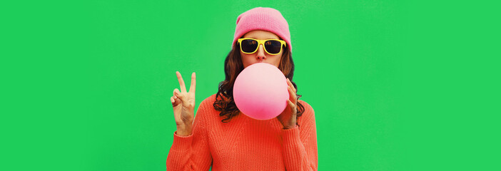 Fashionable portrait of stylish cool young woman inflating chewing gum or balloon wearing pink hat on green background, blank copy space for advertising text