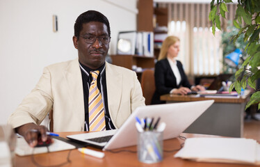 Portrait of adult african american office employee focused on working with laptop..