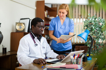 Portrait of two multinational doctors in the office of a resident of the clinic, sitting at a table...