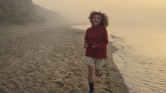 Happy woman running on sea beach at sunrise. Playful girl smiling at camera
