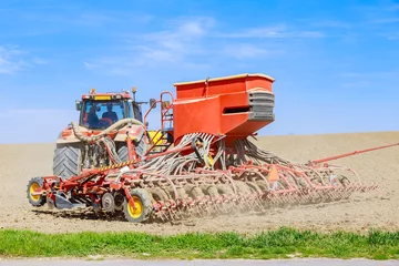 Fotobehang A red tractor sows grain with a seeder in the field. © Yaroslav