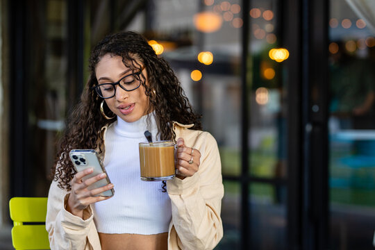 Young Woman Checks Her Phone While Holding A Coffee In A Cafe