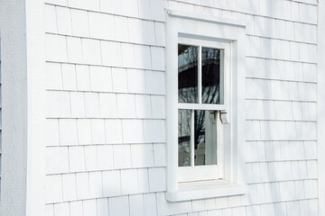 The exterior of a vintage building is covered in white cedar wood shingles. There's a small single hung glass window in the center with the reflection of a white picket fence and tree in the window. 