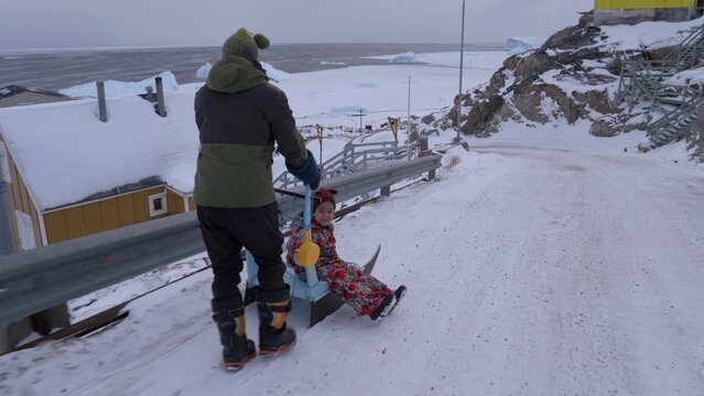 Dad pushes young daughter down a snowy road sitting on sleigh