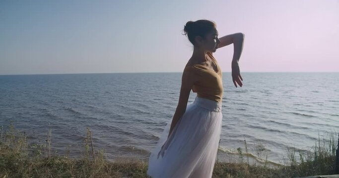Tracking shot side view of slim talented Caucasian woman in tutu walking dancing in slow motion at background of sea river. Medium shot of graceful gorgeous ballerina performing. Cinema 4k ProRes HQ