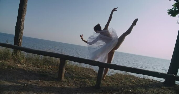 Live camera follows movement of graceful ballerina doing developpe in slow motion at background of river sea. Wide shot Caucasian woman in tutu performing on sunny day outdoors. Cinema 4k ProRes HQ