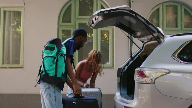 Married couple with luggage going on vacation while loading car trunk with trolley and backpack. Boyfriend and girlfriend getting ready for citybreak departure while putting baggage inside vehicle.