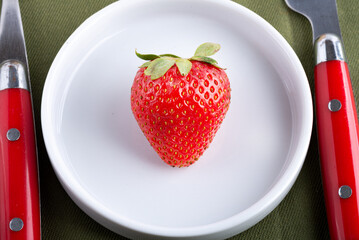 large red strawberry with green leaves in a white porcelain saucer on the dining table. Cutlery.