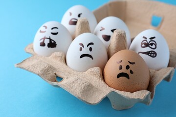 Brown egg with upset face among aggressively disposed white ones in carton box on turquoise...
