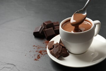 Spoon with yummy hot chocolate above cup on dark table, space for text