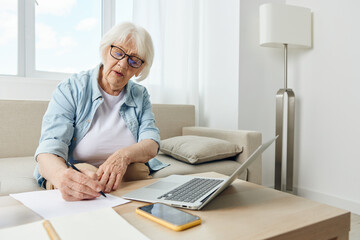 A mature elderly woman in a bright apartment is sitting at a table on the sofa using a laptop. Active retirement lifestyle. The concept of studying leisure.