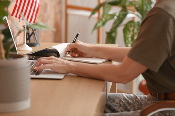 Mature female soldier using laptop and writing in notebook at home