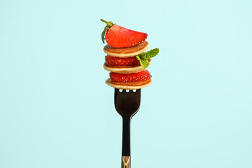 Fork with mini pancakes and fresh strawberry on color background