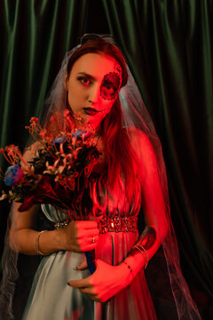 Beautiful girl in a blue dress with artistic makeup on her face holds a bouquet of flowers. Halloween mask. Red light. Halloween concept, nightclub, masquerade