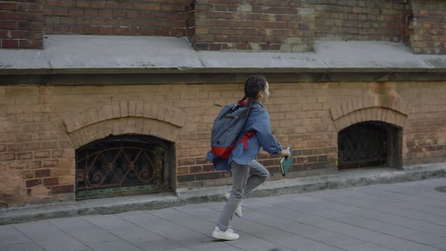 Little girl with a backpack and textbook runs hurrying to school. Little schoolgirl with a backpack runs to the school building. Child running with textbook side view. Education concept