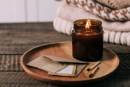 Burning candle in small amber glass jar with wooden wick, stack knitted season sweaters. Cozy lifestyle, hygge concept
