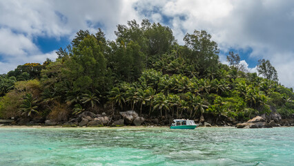 Fototapeta na wymiar A tropical island in the Indian Ocean is overgrown with lush vegetation. Picturesque boulders at the water's edge. The boat is moored at the shore. Blue sky with clouds. Seychelles. Moyenne Island
