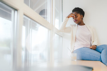 African american woman sitting on the windowsill tired, rubbing nose and eyes feeling fatigue. Depression and sadness. Upset and irritated for problem