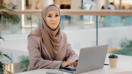 Focused serious arab businesswoman professional entrepreneur sitting at office desk typing looking at laptop screen studying online course muslim girl freelancer works remotely using computer device