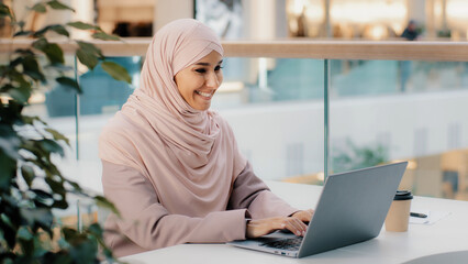 Happy young arab businesswoman sitting at office desk typing on laptop working on internet checking new app smiling looking at camera showing thumb up approval sign consent support symbol high rating