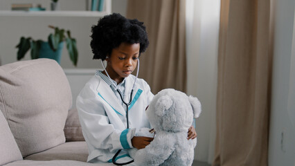 Serious african american kid girl sitting in room on couch in medical uniform pretending to be doctor little child playing veterinarian heals toy patient holding stethoscope listening sick teddy bear