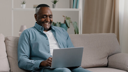 Mature happy african american man sitting on sofa at home relaxing smiling chatting on laptop via webcam talking on video call friendly conference showing thumb up gesture of approval sign agreement