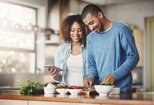 Young, happy and romantic couple cooking healthy food together following recipes online on a tablet. Smiling husband and wife making dinner or lunch in the kitchen at home