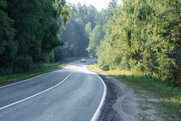 an empty asphalt road in a summer forest with a car in the distance