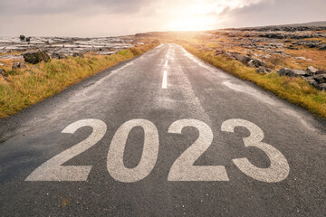 Sign 2023 on a small straight asphalt road at sunrise. New year plans and resolution. Warm color tone. Sun flare and glow.