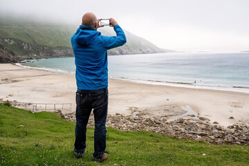 Male tourist taking picture on phone of stunning nature scenery with sandy beach, ocean and...