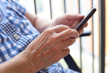 Elderly woman sitting with smartphone in chair, mobile phone in wrinkled female hands closeup. Concept of online communication in retirement