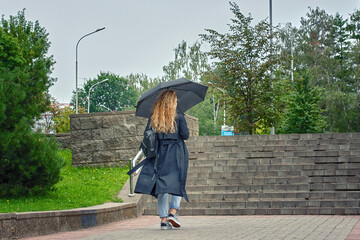 Woman wearing black–coloured trenchcoat while walking the street during light rain. Woman in...