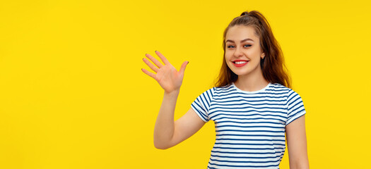 Friendly brunette girl smiling, saying hi and waving hand to greet person, welcome someone,...