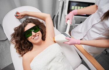 Cosmologist removing hair from female armpit with laser device. Happy woman in protective glasses...