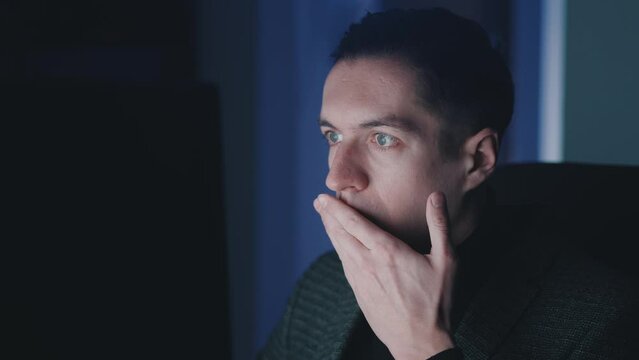 Afraid and shocked man looking at monitor screen, takes off glasses and cover his mouth with his hand while working at computer laptop late at night. Male scared of bad news, read terrible message.
