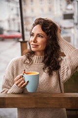 A middle-aged woman in a beige sweater with a blue mug in her hands is in a street cafe on the veranda