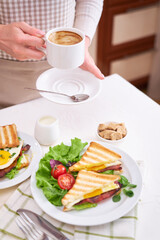 Woman holding cup of fresh espresso coffee over kitchen table with with toasted sandwiches