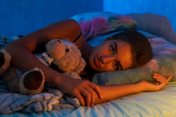 Cute young woman hugging teddy bear, sleeping lying in cozy bed, happy caucasian girl hugging teddy bears, falling asleep on soft pillows under the light of a night lamp