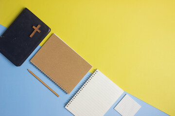 Holy Bible with brown notebook and cross over the yellow and blue background. 