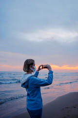 Traveling woman in medial mask taking photo of wonderful sunset on sea beach.