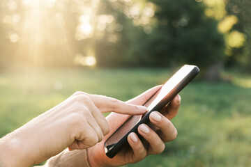 Closeup of female hands using a smart phone, nature background. Woman using mobile phone in sunset outdoors. Unrecognizable person touching smartphone screen. Unknown girl holding cellphone outside