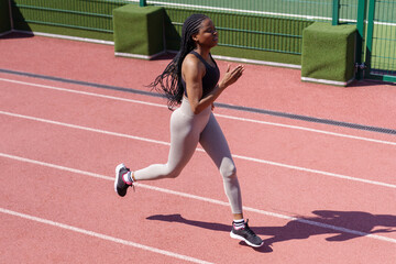 Obraz premium Young black sportswoman enjoys running on red track around stadium to improve technique and increase endurance. African American woman with long braids prepares for upcoming marathon on hot summer day