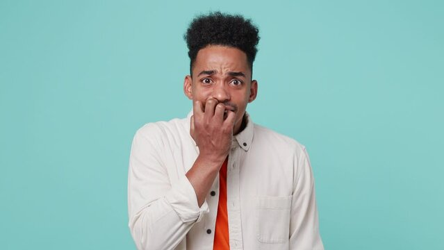 Young scared fearful man of African American ethnicity 20s he wear shirt t-shirt looking camera biting nails fingers isolated on plain pastel light blue cyan color background. People lifestyle concept