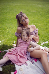 beautiful young mother with a baby daughter in pink dresses sitting on a plaid in a green meadow in nature with a picnic in summer.
